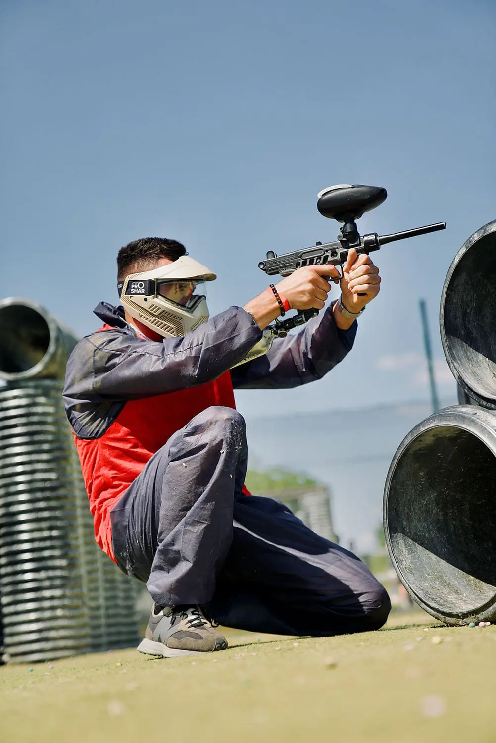 Participant agenouillé derrière des tuyaux, visant avec un lanceur de paintball sur un terrain extérieur