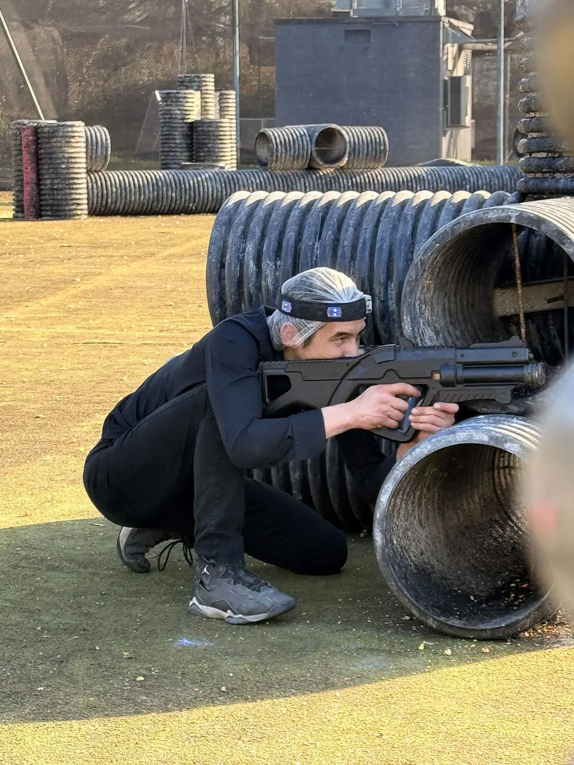 Participant accroupi derrière des tuyaux, visant avec un lanceur de paintball sur un terrain extérieur
