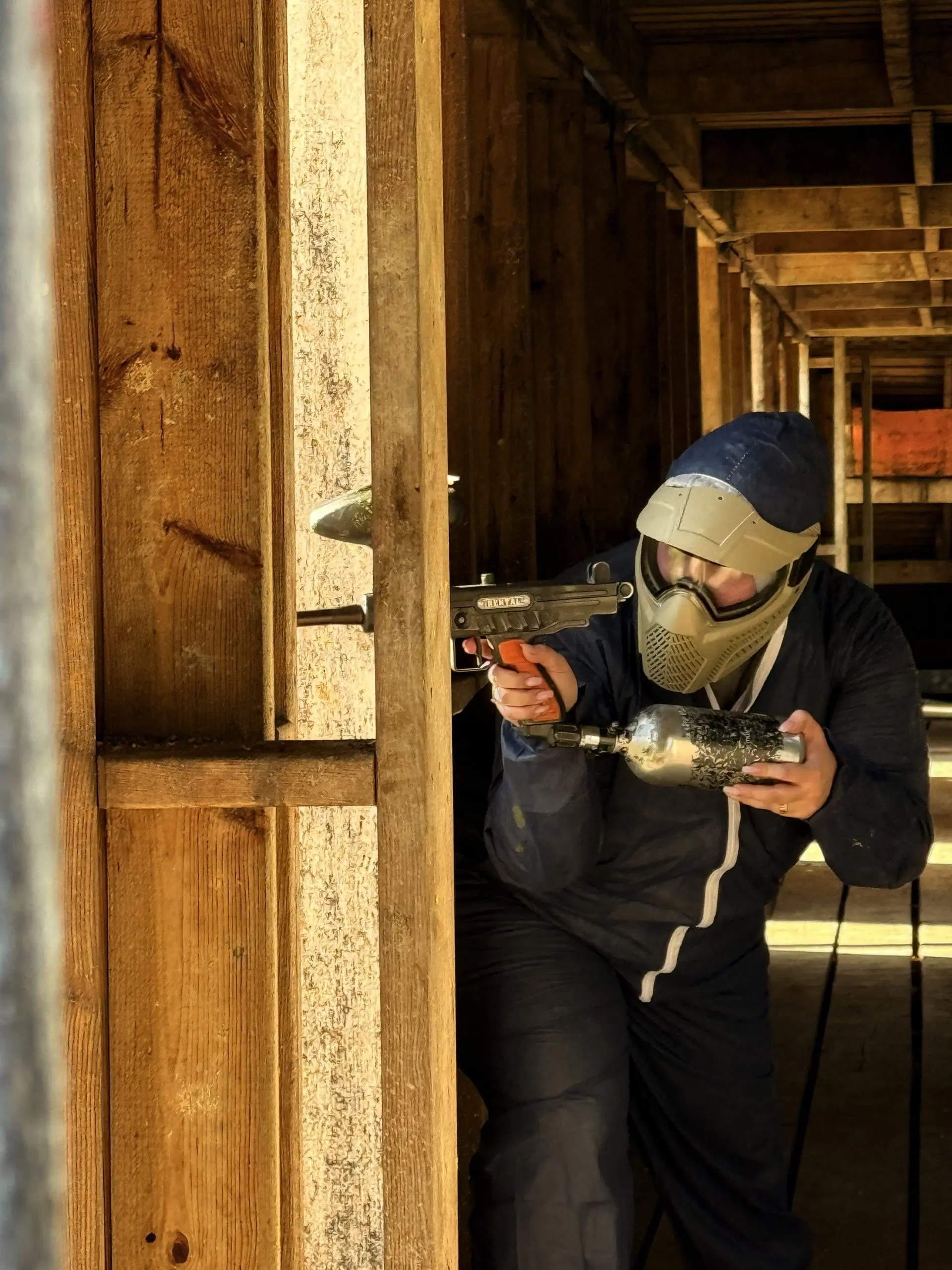 Joueur équipé d’un masque visant avec un lanceur de paintball depuis l’intérieur d’une structure en bois