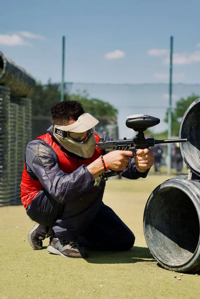Participant agenouillé derrière des tuyaux, visant avec un lanceur de paintball sur un terrain extérieur